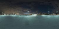 a snow covered parking lot next to tall buildings with lights on it at night with city and sky in the background