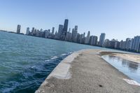 a concrete wall stretches down the side of the ocean towards a city skyline, on a sunny day