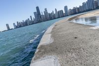 a concrete wall stretches down the side of the ocean towards a city skyline, on a sunny day