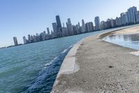 a concrete wall stretches down the side of the ocean towards a city skyline, on a sunny day