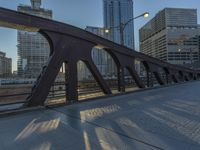 the metal structure on the bridge is long with several crosses in it and there are tall buildings in the background