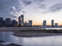 a large city skyline is in the background with a body of water at sunset and beach chairs around it