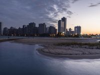 a large city skyline is in the background with a body of water at sunset and beach chairs around it