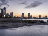 a large city skyline is in the background with a body of water at sunset and beach chairs around it