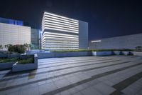 a very tall building with some lights in the background is illuminated from underneath it and sits along an empty floor in a plaza