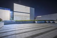 a very tall building with some lights in the background is illuminated from underneath it and sits along an empty floor in a plaza