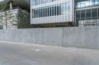 concrete wall next to buildings with glass windows next to a sidewalk in city setting with skateboard on it