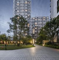 a brick walkway leads to tall buildings in an urban setting at dusk a person is seen standing on the sidewalk, beside trees and bushes