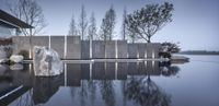 stone sculptures outside a glass building next to the water with trees in the background at dusk