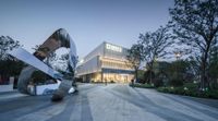 an abstract sculpture near the entrance of a building at dusk with the city light on