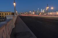 a city bridge at dusk has cars driving along it with buildings lit up in the distance