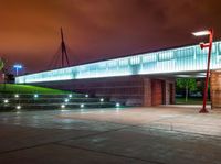 a bridge over a large building with lights on at night time with grass around the walkway and street light fixtures