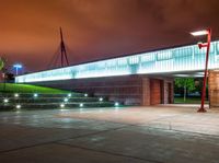 a bridge over a large building with lights on at night time with grass around the walkway and street light fixtures