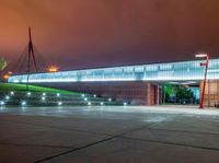 a bridge over a large building with lights on at night time with grass around the walkway and street light fixtures