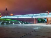 a bridge over a large building with lights on at night time with grass around the walkway and street light fixtures