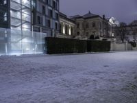 a building with lights shining in the snow in a city area at night time, with snow on ground and light in parking lot