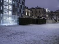 a building with lights shining in the snow in a city area at night time, with snow on ground and light in parking lot