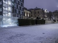 a building with lights shining in the snow in a city area at night time, with snow on ground and light in parking lot