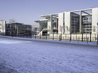 a paved area next to buildings on a clear day with snow on the ground and one side has tall glass windows