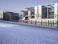 a paved area next to buildings on a clear day with snow on the ground and one side has tall glass windows