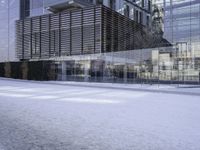 glass building with several buildings next to it in the snow of winter time in front of a tree and some bushes on the sidewalk