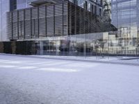 glass building with several buildings next to it in the snow of winter time in front of a tree and some bushes on the sidewalk