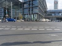 people are walking in a city intersection with a blue car driving past them on the road