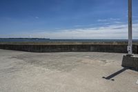 a sidewalk with concrete walls and no clouds in the sky, next to a cement wall that is in the distance there is a man standing on the water's edge of a dock