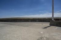 a sidewalk with concrete walls and no clouds in the sky, next to a cement wall that is in the distance there is a man standing on the water's edge of a dock