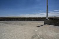 a sidewalk with concrete walls and no clouds in the sky, next to a cement wall that is in the distance there is a man standing on the water's edge of a dock