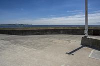 a sidewalk with concrete walls and no clouds in the sky, next to a cement wall that is in the distance there is a man standing on the water's edge of a dock