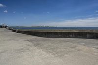 a sidewalk with concrete walls and no clouds in the sky, next to a cement wall that is in the distance there is a man standing on the water's edge of a dock