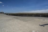 a sidewalk with concrete walls and no clouds in the sky, next to a cement wall that is in the distance there is a man standing on the water's edge of a dock