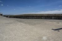 a sidewalk with concrete walls and no clouds in the sky, next to a cement wall that is in the distance there is a man standing on the water's edge of a dock