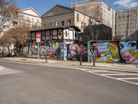 a wall with different colorful artwork on it in a city with buildings around and the street has a bike path going under it