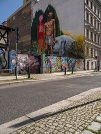 a wall with different colorful artwork on it in a city with buildings around and the street has a bike path going under it