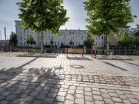 a park bench on the pavement with buildings and trees in the background on a sunny day