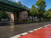 A street with wet pavement reflects light, bordered by a red bike lane. An elevated green metal bridge crosses overhead, supported by stone pillars with graffiti. Trees line the right side, and a pedestrian is visible on the left. A gray car is parked under the bridge.
