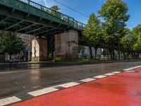 A street with wet pavement reflects light, bordered by a red bike lane. An elevated green metal bridge crosses overhead, supported by stone pillars with graffiti. Trees line the right side, and a pedestrian is visible on the left. A gray car is parked under the bridge.