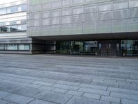 empty plaza with grey stone pavers in front of glass covered building's entrance