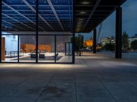 a view outside of a large building through glass windows at dusk with benches and people walking underneath the roof