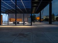 a view outside of a large building through glass windows at dusk with benches and people walking underneath the roof