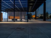 a view outside of a large building through glass windows at dusk with benches and people walking underneath the roof