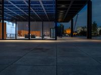 a view outside of a large building through glass windows at dusk with benches and people walking underneath the roof