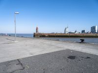 an empty parking lot by the shore with some buildings in the background, and a lighthouse on a small pole