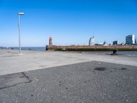 an empty parking lot by the shore with some buildings in the background, and a lighthouse on a small pole