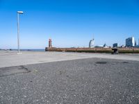 an empty parking lot by the shore with some buildings in the background, and a lighthouse on a small pole