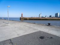 an empty parking lot by the shore with some buildings in the background, and a lighthouse on a small pole