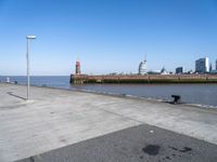 an empty parking lot by the shore with some buildings in the background, and a lighthouse on a small pole