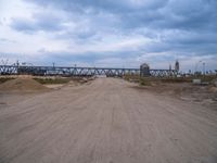 dirt road with train tracks under an overpass on the opposite track, and sand area below with vehicles and machinery